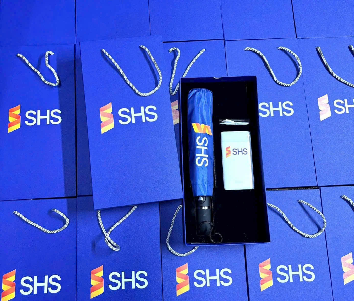 Hộp xanh có dù gấp và vật phẩm chữ nhật, xung quanh là túi xách xanh mang nhận diện SHS