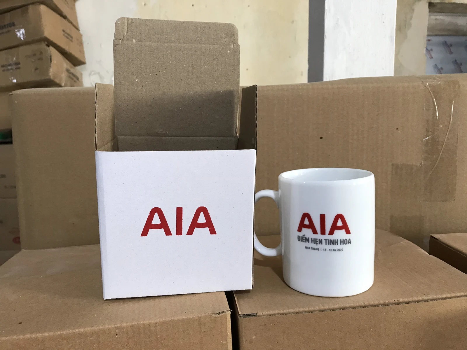 Cốc trắng in AIA cạnh hộp trắng mở nắp, phía sau là các thùng carton
