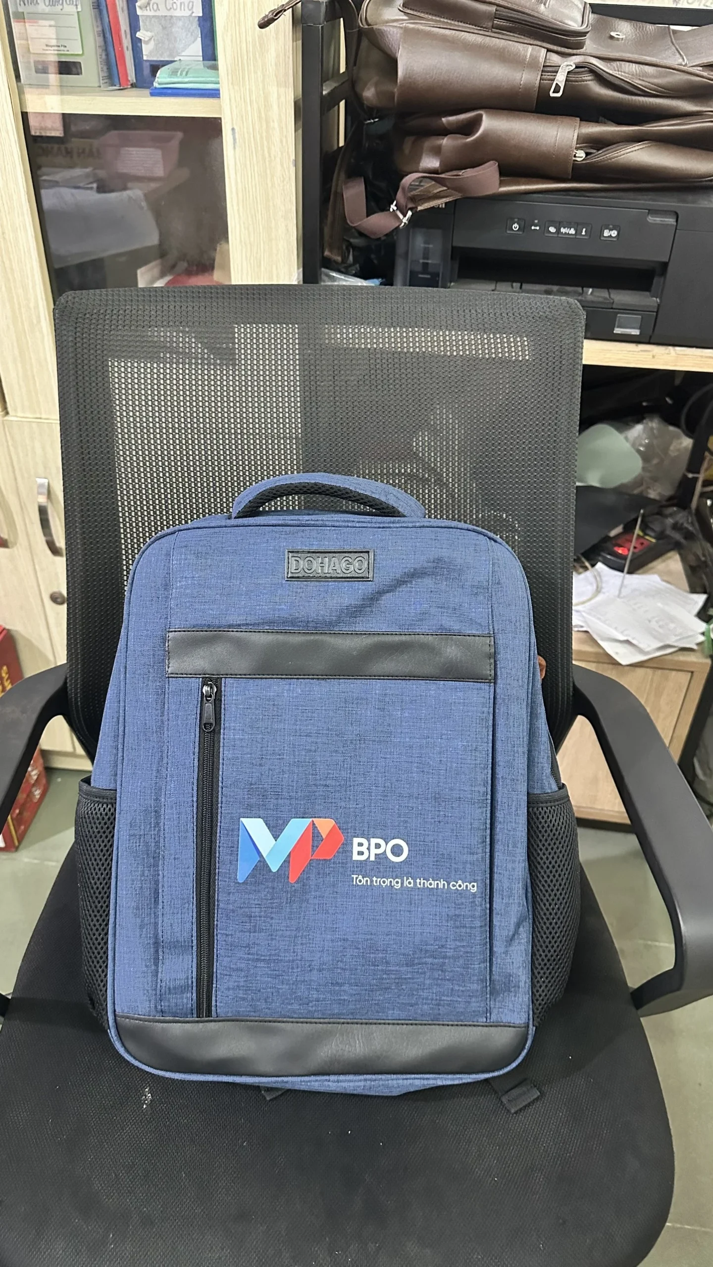 Mặt trước balo xanh có khóa kéo dọc bên trái, nhãn DOHAGO phía trên và logo VP BPO bên dưới