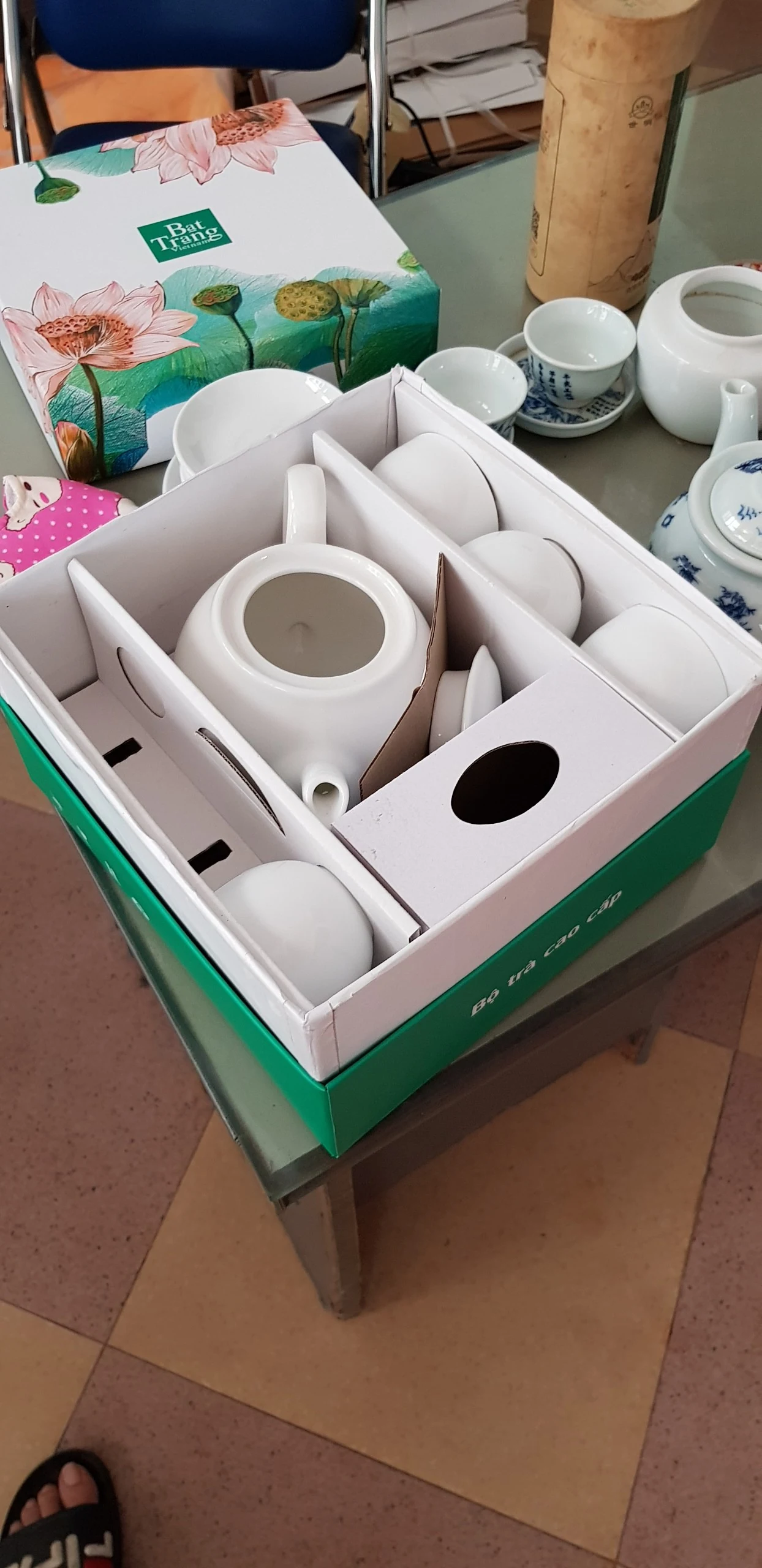 Góc hộp xanh chia vách giấy quanh ấm trắng và các món sứ