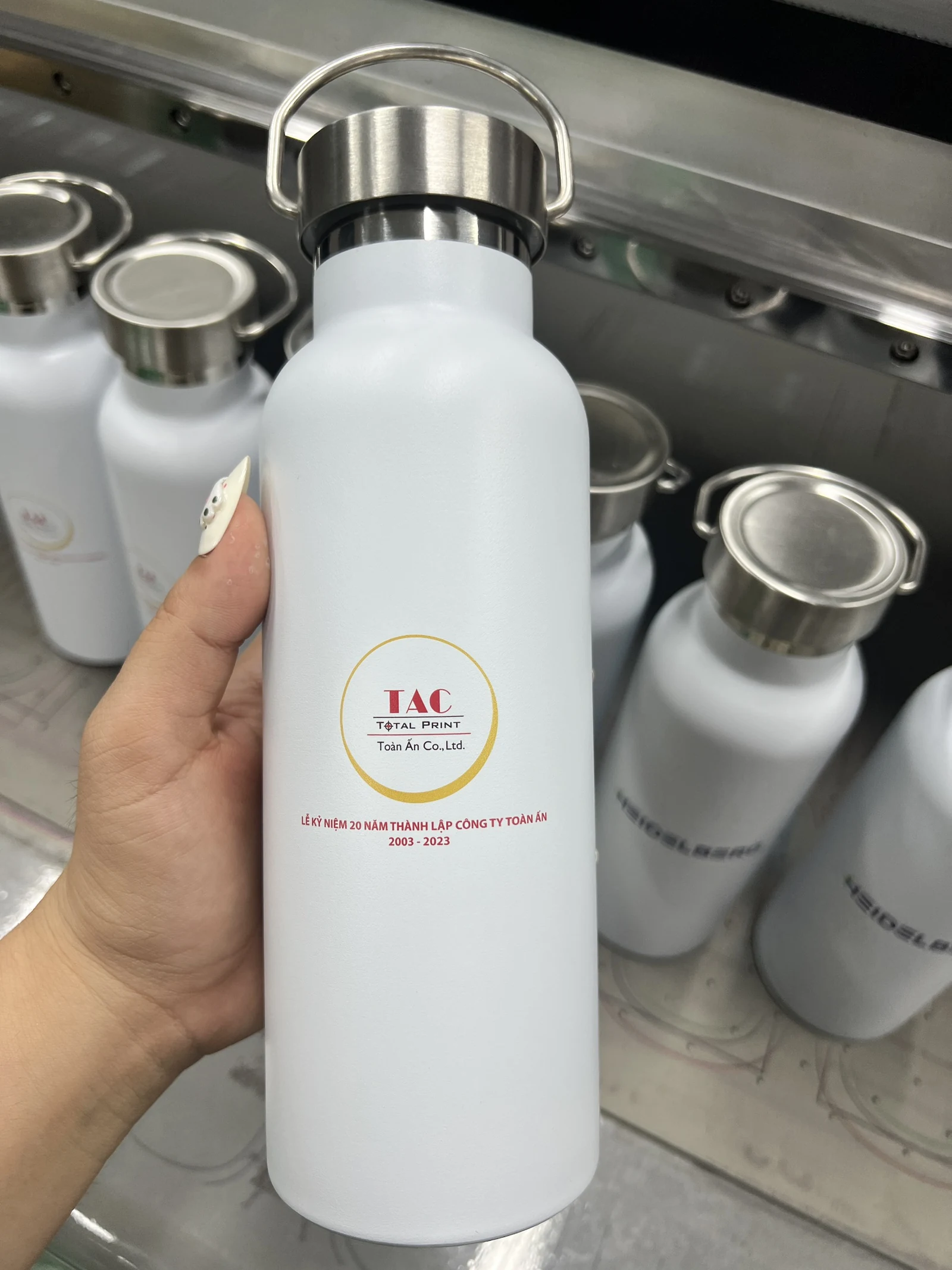 Tay cầm bình trắng nắp bạc có quai, phía sau có bình trắng mở miệng và các bình khác in TAC