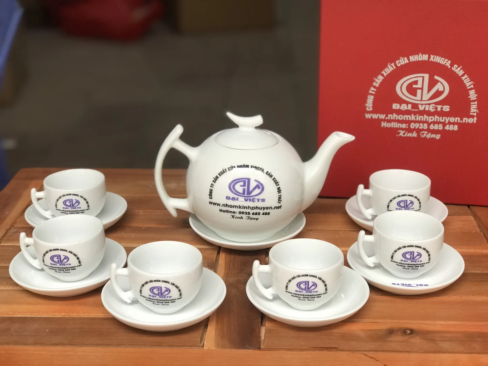 Ảnh bộ trà trắng trên bàn gỗ, chữ logo Đại Việts trên ấm, chén và đĩa, hộp đỏ phía sau
