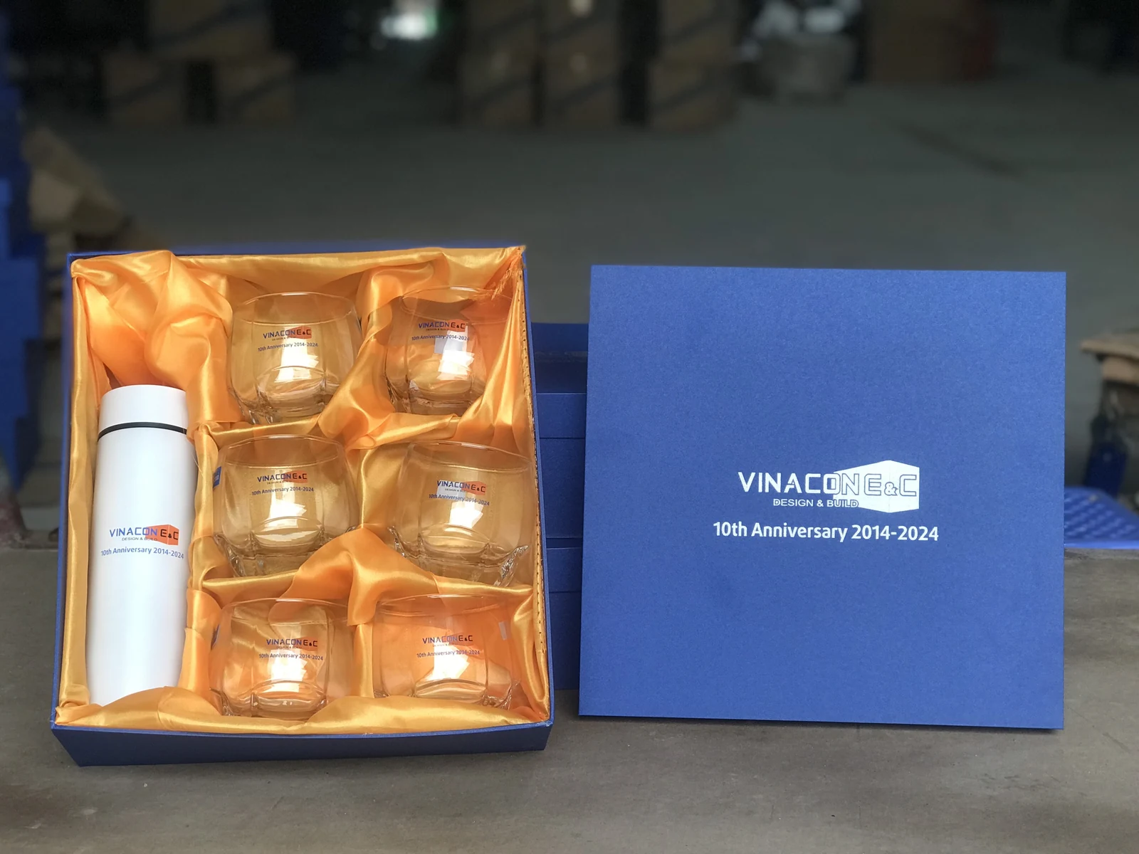 Hộp xanh mở có một bình trắng và sáu ly trong lót vàng, nắp hộp bên phải