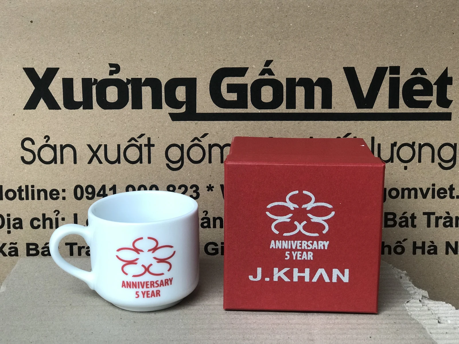 Ảnh chụp cốc trắng có biểu tượng và chữ đỏ cạnh hộp đỏ có biểu tượng trắng, phía sau là bảng Xưởng Gốm Việt