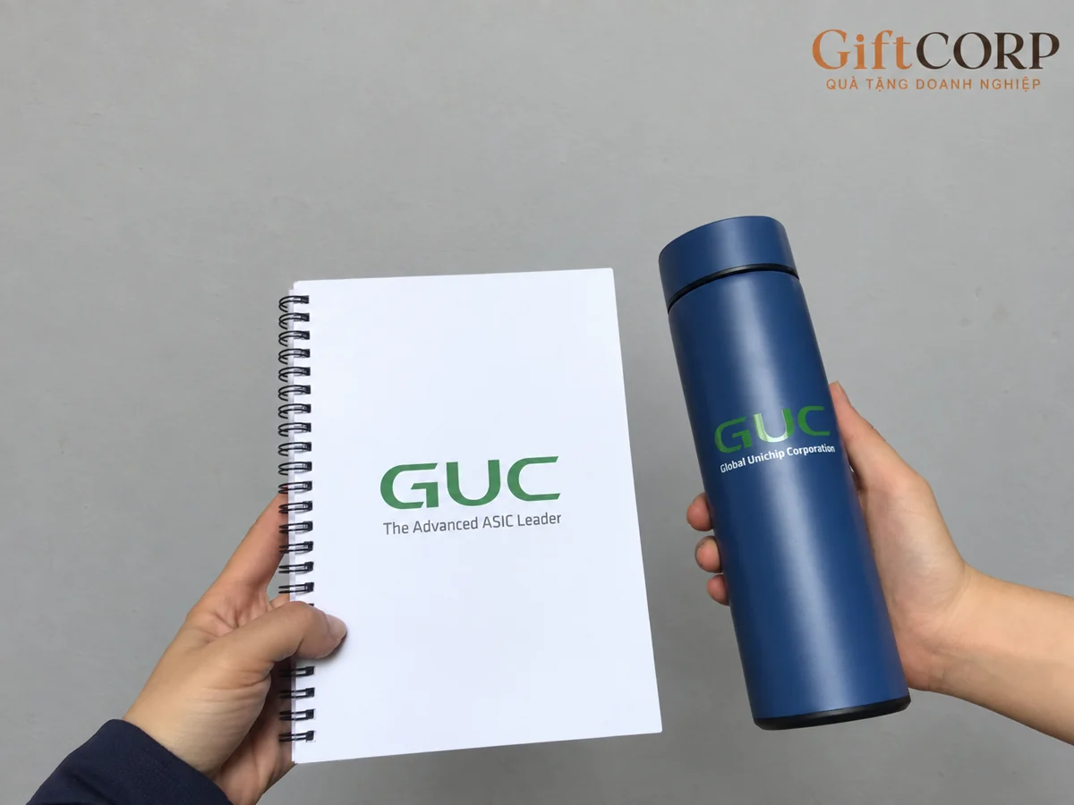 Tay cầm sổ trắng lò xo và bình xanh đậm in GUC, hậu cảnh xám có logo GiftCorp